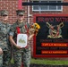 Two female U.S. Marines in MARPAT camouflage uniforms pose together in front of a 'Bravo Nation' company sign. One Marine is holding a bouquet of flowers and a framed certificate with a red ribbon, suggesting a recognition or promotion ceremony. The sign identifies the unit's Company Commander as Capt A.P. McCollum and Company First Sergeant as 1stSgt K.F. Martinezlopez.
