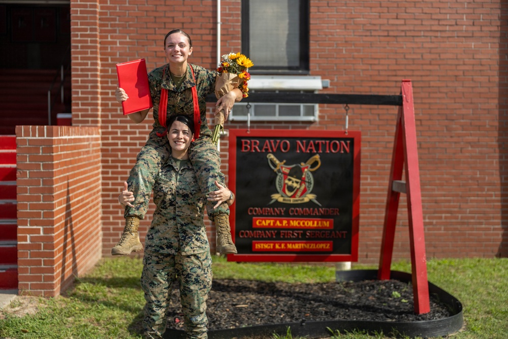 Two female U.S. Marines in woodland MARPAT camouflage uniforms pose playfully in front of a 'Bravo Nation' company sign. One Marine is sitting on the shoulders of the other, holding a red folder and a bouquet of sunflowers, suggesting a celebratory occasion such as a promotion or award ceremony. The sign behind them identifies the unit's Company Commander as Capt. A.P. McCollum and Company First Sergeant as 1stSgt K.E. MartinezLopez.