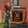 Two female U.S. Marines in woodland MARPAT camouflage uniforms pose playfully in front of a 'Bravo Nation' company sign. One Marine is sitting on the shoulders of the other, holding a red folder and a bouquet of sunflowers, suggesting a celebratory occasion such as a promotion or award ceremony. The sign behind them identifies the unit's Company Commander as Capt. A.P. McCollum and Company First Sergeant as 1stSgt K.E. MartinezLopez.