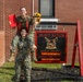 Two female U.S. Marines in woodland MARPAT camouflage uniforms pose playfully in front of a 'Bravo Nation' company sign. One Marine is sitting on the shoulders of the other, holding a red folder and a bouquet of sunflowers, suggesting a celebratory occasion such as a promotion or award ceremony. The sign behind them identifies the unit's Company Commander as Capt. A.P. McCollum and Company First Sergeant as 1stSgt K.E. MartinezLopez.