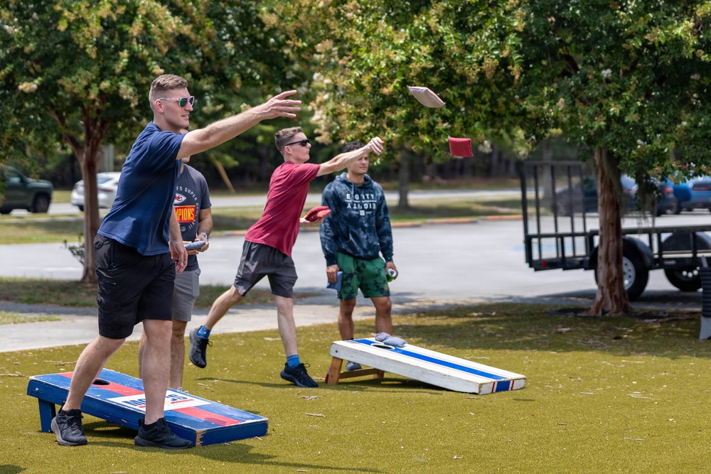 A group of young men are playing cornhole (bean bag toss) outdoors on a grass or turf surface. Two players in the foreground are actively throwing bean bags toward the opposite boards, while a third player stands in the background observing. The setting appears to be a casual outdoor recreational area with trees, parked vehicles, and a utility trailer visible in the background.