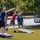 MCAS Beaufort Summer Bash A group of young men are playing cornhole (bean bag toss) outdoors on a grass or turf surface. Two players in the foreground are actively throwing bean bags toward the opposite boards, while a third player stands in the background observing. The setting appears to be a casual outdoor recreational area with trees, parked vehicles, and a utility trailer visible in the background.