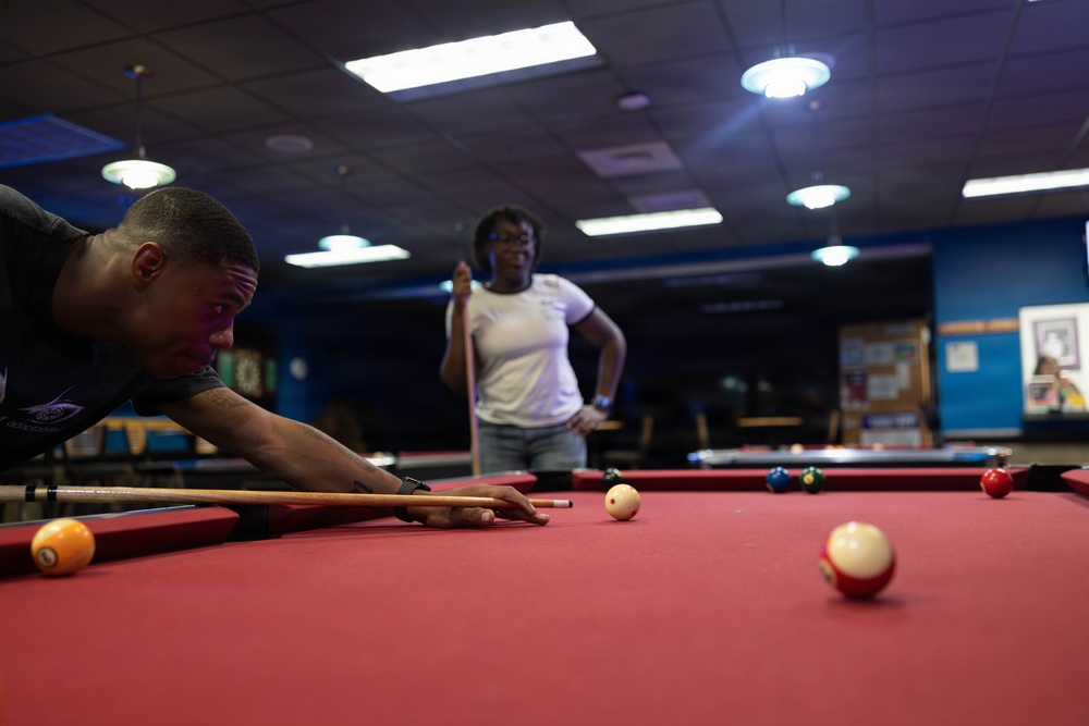 Two individuals are playing pool/billiards in what appears to be a recreational billiards hall or game room. A man is leaning over the red-felt pool table lining up a shot with his cue stick, while a woman stands in the background holding her cue stick and watching. The room has multiple pool tables, overhead lighting, and blue-painted walls.
