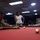 MCAS Beaufort Summer Bash Two individuals are playing pool/billiards in what appears to be a recreational billiards hall or game room. A man is leaning over the red-felt pool table lining up a shot with his cue stick, while a woman stands in the background holding her cue stick and watching. The room has multiple pool tables, overhead lighting, and blue-painted walls.