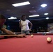 Two individuals are playing pool/billiards in what appears to be a recreational billiards hall or game room. A man is leaning over the red-felt pool table lining up a shot with his cue stick, while a woman stands in the background holding her cue stick and watching. The room has multiple pool tables, overhead lighting, and blue-painted walls.