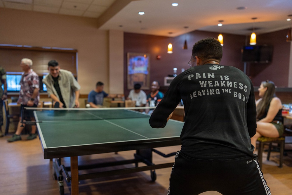 A group of people are gathered in what appears to be a recreational room or lounge, with the foreground showing a person playing ping pong. The player in the foreground wears a black long-sleeve shirt reading 'PAIN IS WEAKNESS LEAVING THE BODY' with a Marine Corps emblem, suggesting a military or veteran-affiliated setting. Several other individuals are visible in the background, either watching, socializing, or playing at other tables.