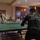 MCAS Beaufort Summer Bash A group of people are gathered in what appears to be a recreational room or lounge, with the foreground showing a person playing ping pong. The player in the foreground wears a black long-sleeve shirt reading 'PAIN IS WEAKNESS LEAVING THE BODY' with a Marine Corps emblem, suggesting a military or veteran-affiliated setting. Several other individuals are visible in the background, either watching, socializing, or playing at other tables.