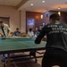 A group of people are gathered in what appears to be a recreational room or lounge, with the foreground showing a person playing ping pong. The player in the foreground wears a black long-sleeve shirt reading 'PAIN IS WEAKNESS LEAVING THE BODY' with a Marine Corps emblem, suggesting a military or veteran-affiliated setting. Several other individuals are visible in the background, either watching, socializing, or playing at other tables.