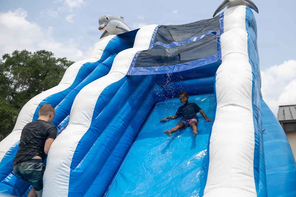Two children are playing on a large inflatable water slide decorated with dolphin/shark motifs in blue and white colors. One child is actively sliding down the wet surface while another child is climbing up the side. The setting appears to be an outdoor event on a sunny, partly cloudy day.