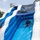MCAS Beaufort Summer Bash Two children are playing on a large inflatable water slide decorated with dolphin/shark motifs in blue and white colors. One child is actively sliding down the wet surface while another child is climbing up the side. The setting appears to be an outdoor event on a sunny, partly cloudy day.
