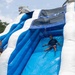Two children are playing on a large inflatable water slide decorated with dolphin/shark motifs in blue and white colors. One child is actively sliding down the wet surface while another child is climbing up the side. The setting appears to be an outdoor event on a sunny, partly cloudy day.