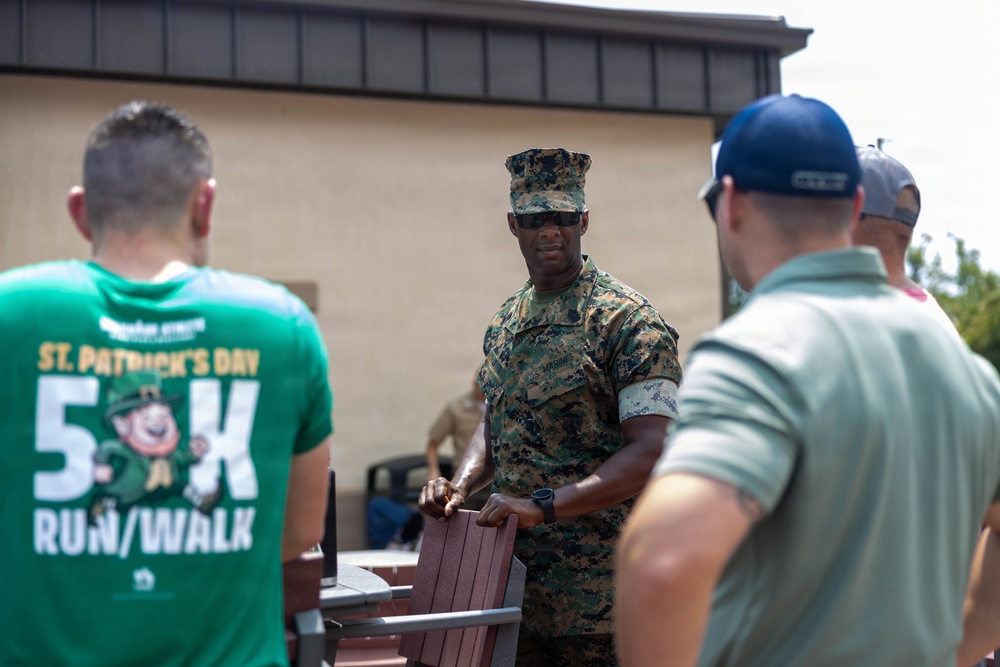 A U.S. Marine in MARPAT camouflage uniform and cover is interacting with two civilians outdoors near a building. The Marine appears to be handling or moving a wooden/composite chair. One civilian is wearing a green St. Patrick's Day 5K Run/Walk event t-shirt.