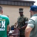 A U.S. Marine in MARPAT camouflage uniform and cover is interacting with two civilians outdoors near a building. The Marine appears to be handling or moving a wooden/composite chair. One civilian is wearing a green St. Patrick's Day 5K Run/Walk event t-shirt.