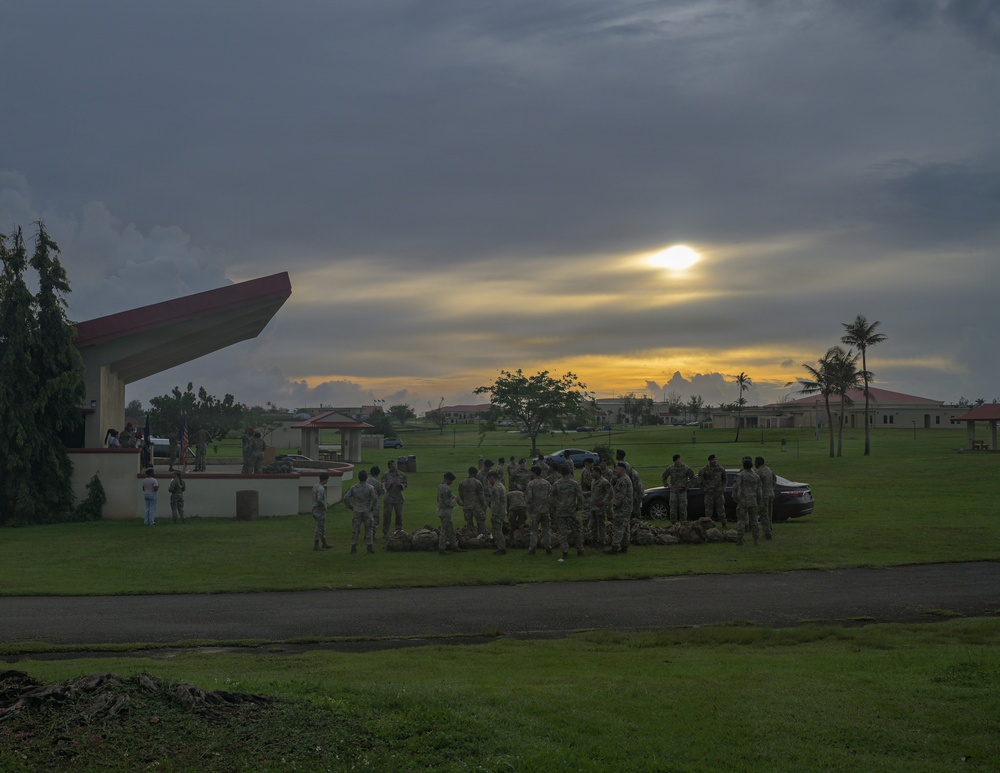 A group of military personnel in camouflage uniforms are gathered on a grassy area at what appears to be a military installation during sunset or sunrise. A covered pavilion or memorial structure with a red roof is visible on the left, with what appears to be an American flag. The dramatic sky features dark storm clouds with golden light breaking through, and palm trees and low-rise buildings are visible in the background.