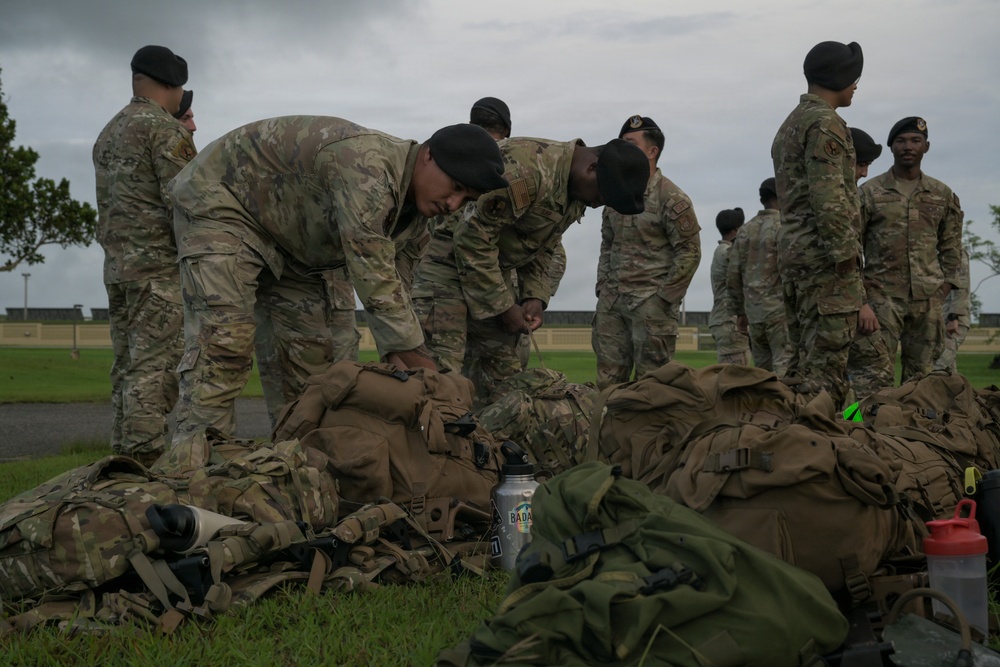 A group of military personnel in OCP (Operational Camouflage Pattern) uniforms and black berets are gathered outdoors on a grassy area. Two soldiers in the foreground are leaning over and inspecting or organizing a collection of tactical rucksacks and gear laid out on the ground. The overcast sky and military installation buildings visible in the background suggest a field training or pre-deployment equipment inspection activity.