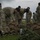 Team Andersen kick-off 24-hour POW/MIA memorial event A group of military personnel in OCP (Operational Camouflage Pattern) uniforms and black berets are gathered outdoors on a grassy area. Two soldiers in the foreground are leaning over and inspecting or organizing a collection of tactical rucksacks and gear laid out on the ground. The overcast sky and military installation buildings visible in the background suggest a field training or pre-deployment equipment inspection activity.