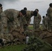 A group of military personnel in OCP (Operational Camouflage Pattern) uniforms and black berets are gathered outdoors on a grassy area. Two soldiers in the foreground are leaning over and inspecting or organizing a collection of tactical rucksacks and gear laid out on the ground. The overcast sky and military installation buildings visible in the background suggest a field training or pre-deployment equipment inspection activity.