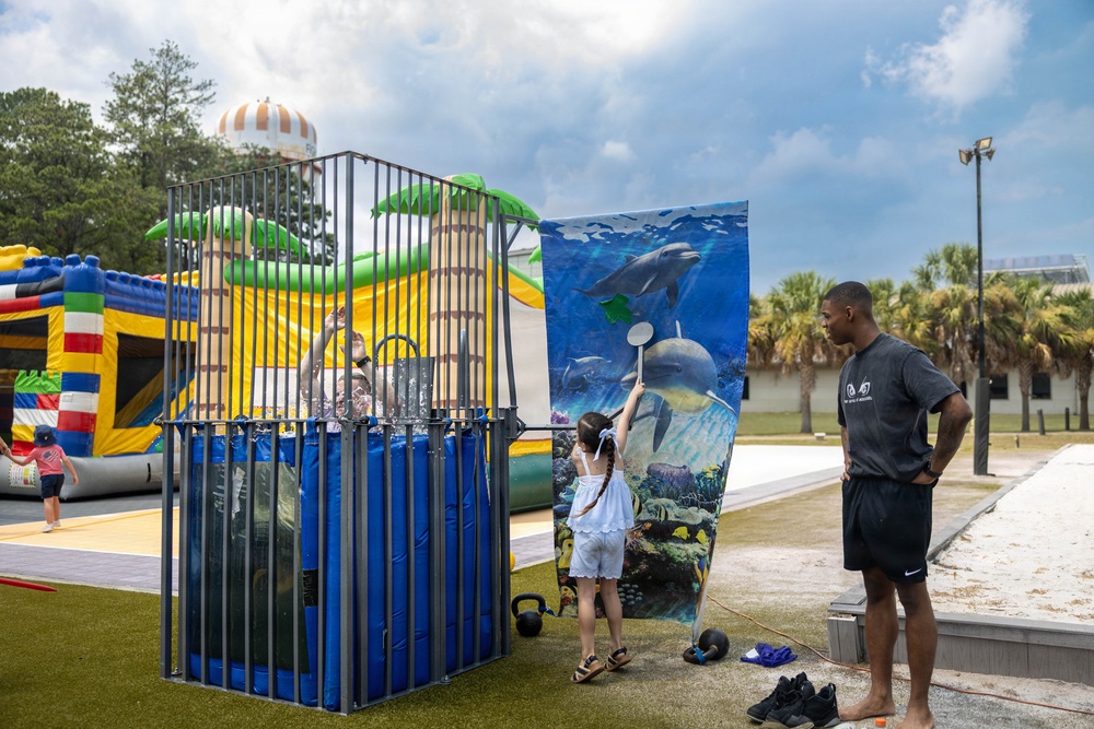 An outdoor carnival or community event featuring a dunk tank where a person is seated inside getting splashed with water. A young girl is attempting to trigger the dunk tank mechanism while an adult male supervises nearby. The setting includes inflatable bounce houses, palm trees, and a paved recreational area.