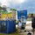 MCAS Beaufort Summer Bash An outdoor carnival or community event featuring a dunk tank where a person is seated inside getting splashed with water. A young girl is attempting to trigger the dunk tank mechanism while an adult male supervises nearby. The setting includes inflatable bounce houses, palm trees, and a paved recreational area.