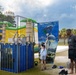 An outdoor carnival or community event featuring a dunk tank where a person is seated inside getting splashed with water. A young girl is attempting to trigger the dunk tank mechanism while an adult male supervises nearby. The setting includes inflatable bounce houses, palm trees, and a paved recreational area.