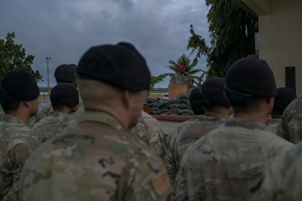 A military formation is gathered outdoors listening to a speaker at a podium, with sandbags stacked in front of the speaking area. The personnel are wearing camouflage uniforms and black berets, suggesting a formal military ceremony or briefing. The overcast sky and tropical vegetation in the background indicate a humid, possibly coastal or island location.