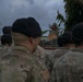 A military formation is gathered outdoors listening to a speaker at a podium, with sandbags stacked in front of the speaking area. The personnel are wearing camouflage uniforms and black berets, suggesting a formal military ceremony or briefing. The overcast sky and tropical vegetation in the background indicate a humid, possibly coastal or island location.