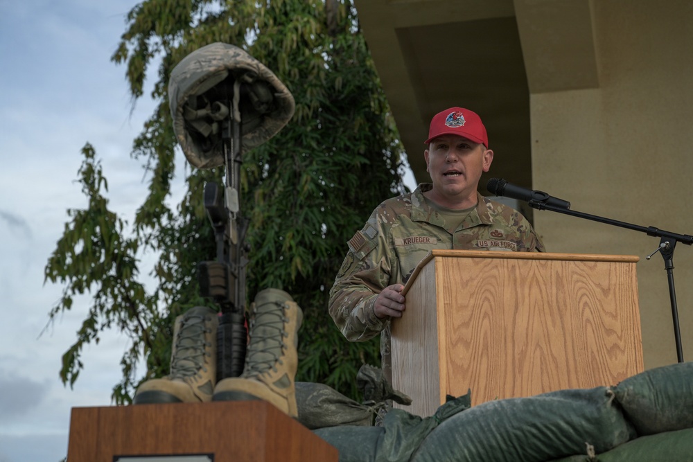 A U.S. Air Force servicemember named Krueger is speaking at a podium during what appears to be a memorial ceremony. Behind him stands a 'Battlefield Cross' or 'Fallen Soldier Battle Cross' — a symbolic tribute consisting of boots, a rifle, and a helmet. The setting appears to be an outdoor military ceremony.