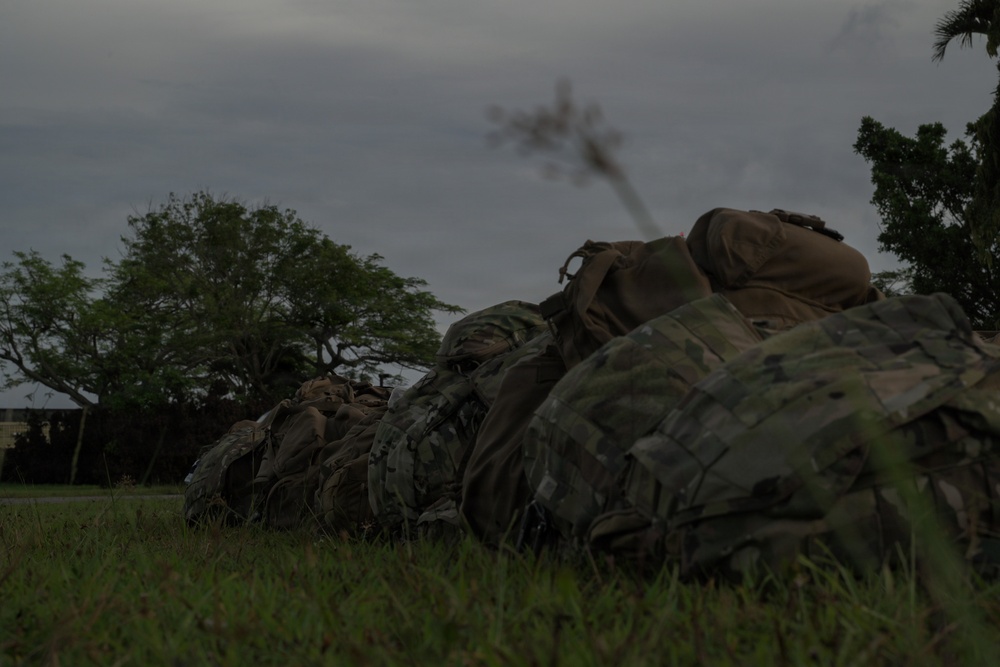 The image shows a row of military rucksacks/backpacks in multicam/OCP camouflage pattern laid out on a grassy field at low light conditions, likely dusk or dawn. The packs appear to be staged in a line, suggesting a military formation or pre-mission equipment staging area. The environment appears tropical or subtropical based on the vegetation visible in the background.