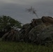The image shows a row of military rucksacks/backpacks in multicam/OCP camouflage pattern laid out on a grassy field at low light conditions, likely dusk or dawn. The packs appear to be staged in a line, suggesting a military formation or pre-mission equipment staging area. The environment appears tropical or subtropical based on the vegetation visible in the background.