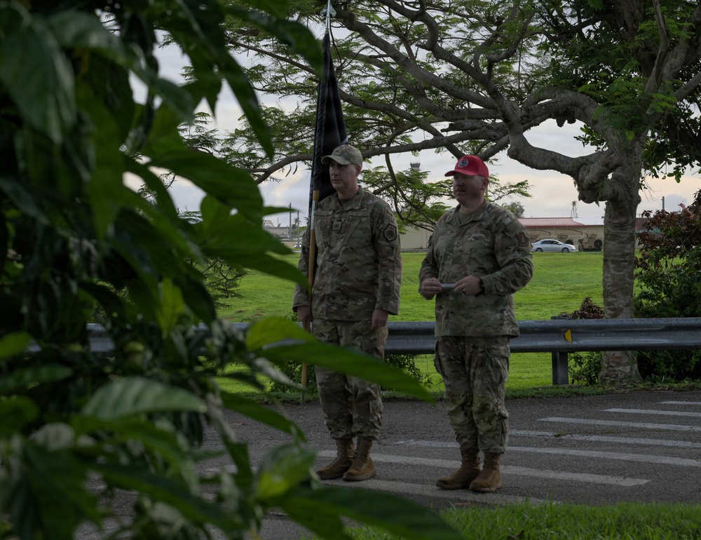 Two military personnel in camouflage uniforms are standing on a paved path in an outdoor setting, likely on a military installation. One soldier holds a dark unit guidon or flag, while the other wears a distinctive red cap and appears to be holding a card or paper. The scene is partially obscured by foreground foliage, suggesting a ceremonial or formal outdoor event.