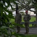 Two military personnel in camouflage uniforms are standing on a paved path in an outdoor setting, likely on a military installation. One soldier holds a dark unit guidon or flag, while the other wears a distinctive red cap and appears to be holding a card or paper. The scene is partially obscured by foreground foliage, suggesting a ceremonial or formal outdoor event.