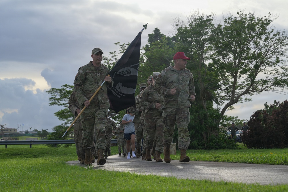 A group of military personnel in camouflage uniforms are marching or running along a paved path in what appears to be a military base or installation. The lead soldier is carrying a black POW/MIA flag on a wooden staff. The setting features lush green grass, tropical trees, and an overcast sky suggesting a warm, humid climate.