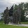 Team Andersen kick-off 24-hour POW/MIA memorial event A group of military personnel in camouflage uniforms are marching or running along a paved path in what appears to be a military base or installation. The lead soldier is carrying a black POW/MIA flag on a wooden staff. The setting features lush green grass, tropical trees, and an overcast sky suggesting a warm, humid climate.