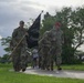 A group of military personnel in camouflage uniforms are marching or running along a paved path in what appears to be a military base or installation. The lead soldier is carrying a black POW/MIA flag on a wooden staff. The setting features lush green grass, tropical trees, and an overcast sky suggesting a warm, humid climate.