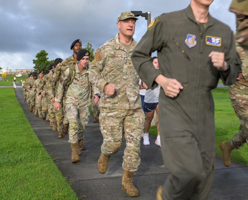 A group of military personnel are participating in a formation run or ruck march along a paved path on what appears to be a military installation. The group includes individuals in various uniforms including OCP camouflage, a green flight suit, and black berets, with some carrying rucksacks. The setting appears to be a tropical or subtropical location based on the lush green grass and overcast sky.