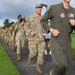 A group of military personnel are participating in a formation run or ruck march along a paved path on what appears to be a military installation. The group includes individuals in various uniforms including OCP camouflage, a green flight suit, and black berets, with some carrying rucksacks. The setting appears to be a tropical or subtropical location based on the lush green grass and overcast sky.