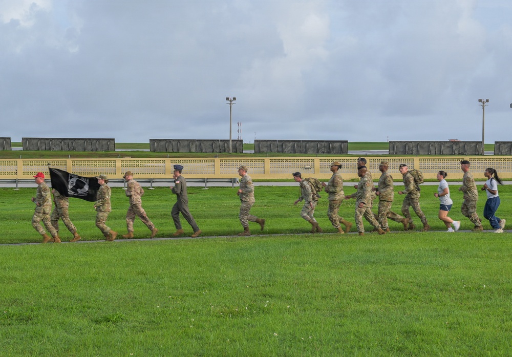 A group of military personnel in camouflage uniforms are running or marching along a paved path on a grassy field, likely on a military installation. The lead individuals are carrying a black POW/MIA flag. Two civilians in casual clothing are also participating at the rear of the group.