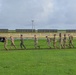 A group of military personnel in camouflage uniforms are running or marching along a paved path on a grassy field, likely on a military installation. The lead individuals are carrying a black POW/MIA flag. Two civilians in casual clothing are also participating at the rear of the group.