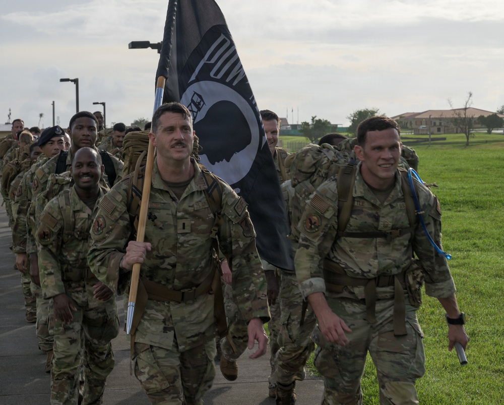 A group of military personnel in camouflage uniforms are conducting a ruck march along a paved path on what appears to be a military installation. The lead soldier is carrying a POW/MIA flag, indicating this is likely a commemorative or memorial ruck march event. The soldiers are wearing full gear including rucksacks and appear to be in good spirits.