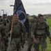 A group of military personnel in camouflage uniforms are conducting a ruck march along a paved path on what appears to be a military installation. The lead soldier is carrying a POW/MIA flag, indicating this is likely a commemorative or memorial ruck march event. The soldiers are wearing full gear including rucksacks and appear to be in good spirits.