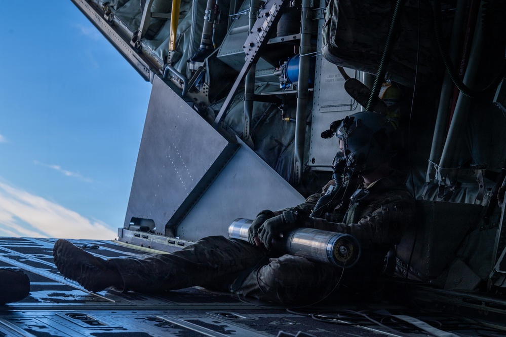 A military aircrew member is seated on the open rear ramp of a large military transport aircraft, likely a C-130 Hercules or similar platform, in flight. The individual is wearing full flight gear including a helmet with oxygen mask and is holding a cylindrical metallic object, possibly a sonobuoy or flare canister. The image is taken from the ramp level looking outward toward a blue sky with light cloud cover.