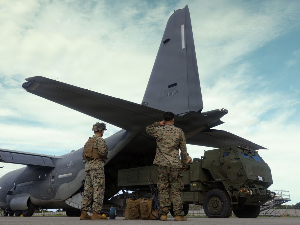 Two U.S. Marines in MARPAT camouflage uniforms stand at the rear cargo ramp of a military transport aircraft, with a green tactical military vehicle positioned alongside. The aircraft tail number '86205' is visible on the vertical stabilizer. The scene appears to depict a loading or unloading operation on a tarmac.