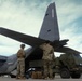 Two U.S. Marines in MARPAT camouflage uniforms stand at the rear cargo ramp of a military transport aircraft, with a green tactical military vehicle positioned alongside. The aircraft tail number '86205' is visible on the vertical stabilizer. The scene appears to depict a loading or unloading operation on a tarmac.