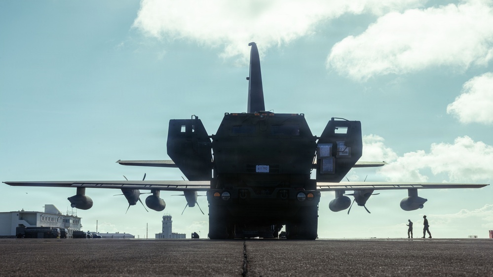 A large military transport aircraft, consistent with a C-130 Hercules, is photographed from the rear on a tarmac with a heavy military vehicle — appearing to be an M142 HIMARS or similar rocket artillery system — positioned directly behind or being loaded into it. The image is taken in strong backlight (contre-jour), rendering both the aircraft and vehicle largely in silhouette. Two personnel are visible in the background near the right side of the aircraft.