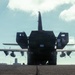 A large military transport aircraft, consistent with a C-130 Hercules, is photographed from the rear on a tarmac with a heavy military vehicle — appearing to be an M142 HIMARS or similar rocket artillery system — positioned directly behind or being loaded into it. The image is taken in strong backlight (contre-jour), rendering both the aircraft and vehicle largely in silhouette. Two personnel are visible in the background near the right side of the aircraft.