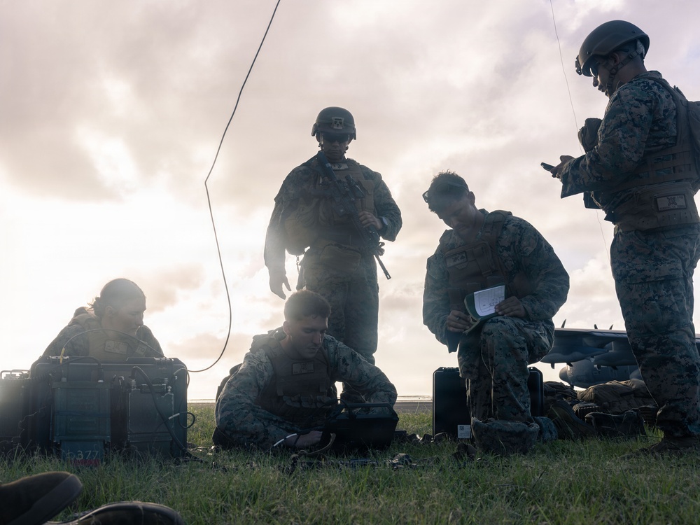 A group of five military personnel in camouflage uniforms are gathered on a grassy field, working with communications or electronic equipment. The scene appears to be a field operation or training exercise, with personnel both kneeling and standing around portable equipment cases and devices. An antenna wire is visibly extended upward, suggesting radio or communications setup activity.