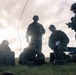 A group of five military personnel in camouflage uniforms are gathered on a grassy field, working with communications or electronic equipment. The scene appears to be a field operation or training exercise, with personnel both kneeling and standing around portable equipment cases and devices. An antenna wire is visibly extended upward, suggesting radio or communications setup activity.