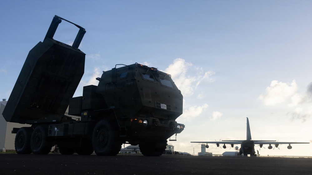The image shows a military rocket artillery system, consistent with an M142 HIMARS (High Mobility Artillery Rocket System), positioned on what appears to be an airfield tarmac. In the background, a large military transport aircraft, consistent with a C-130 Hercules, is visible on the runway. The scene is captured in low-light/silhouette conditions suggesting early morning or dusk.