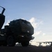 The image shows a military rocket artillery system, consistent with an M142 HIMARS (High Mobility Artillery Rocket System), positioned on what appears to be an airfield tarmac. In the background, a large military transport aircraft, consistent with a C-130 Hercules, is visible on the runway. The scene is captured in low-light/silhouette conditions suggesting early morning or dusk.