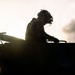 A silhouetted military personnel is positioned atop a large armored or military vehicle, photographed against a dramatic backlit sky with clouds. The strong backlighting creates a high-contrast silhouette effect, obscuring fine details but clearly showing the soldier's camouflage helmet with foliage and combat gear. The composition emphasizes the dramatic, atmospheric quality of the scene.